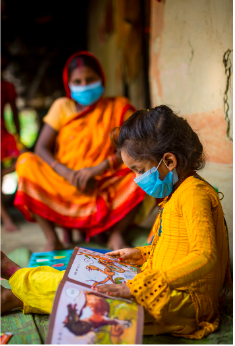 A child in a bright yellow shirt look at a book while her mother watches. Both are wearing blue masks.