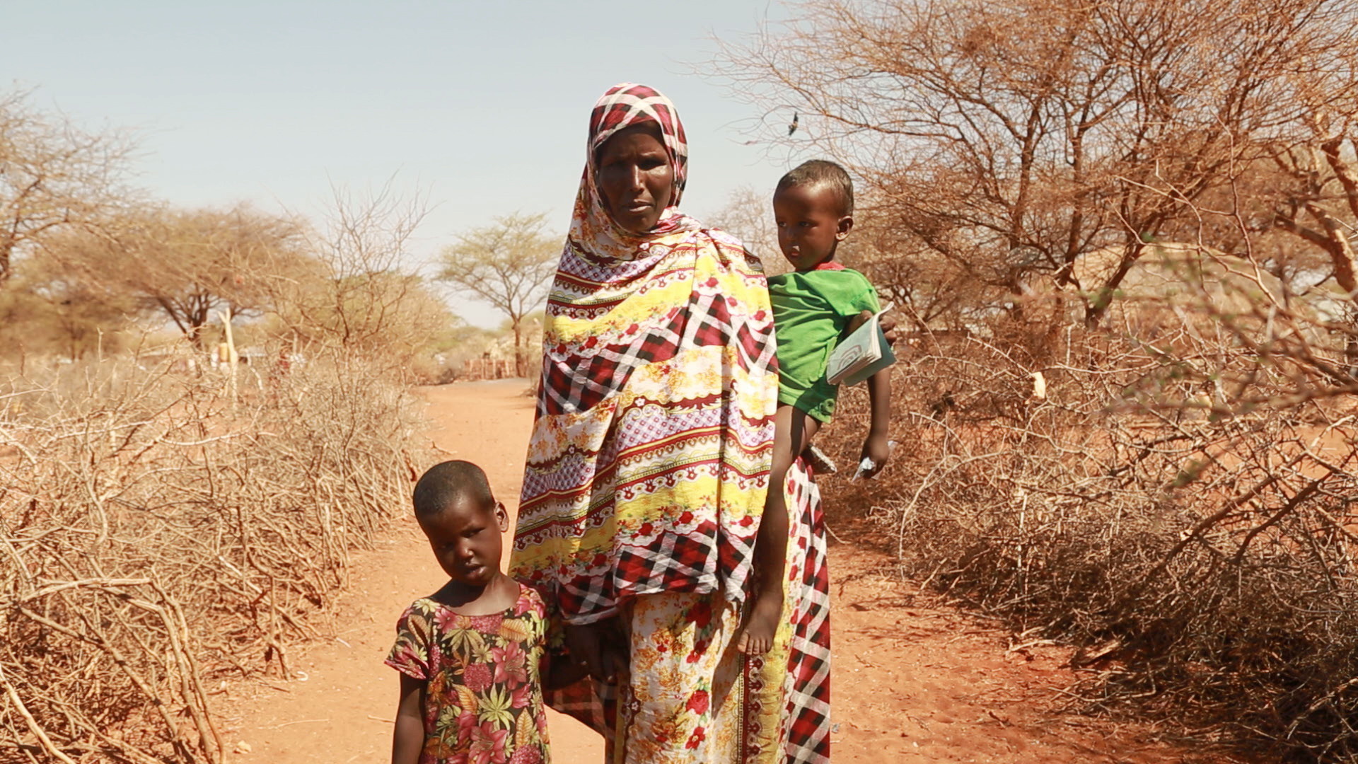 Mother with two young children stand against backdrop of dried out trees and bushes in Kenya